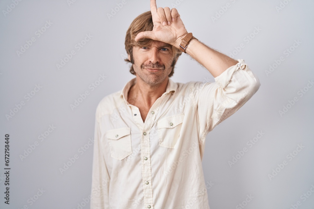 Sticker middle age man standing over isolated background making fun of people with fingers on forehead doing