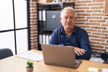Senior man with grey hair working using computer laptop at the office with serious expression on face. simple and natural looking at the camera.