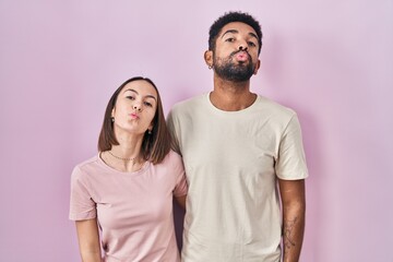 Young hispanic couple together over pink background looking at the camera blowing a kiss on air being lovely and sexy. love expression.