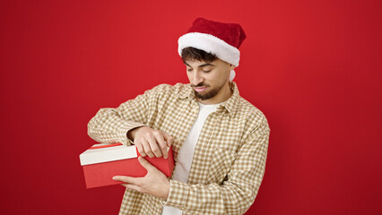 Young arab man wearing christmas hat unpacking gift looking upset over isolated red background