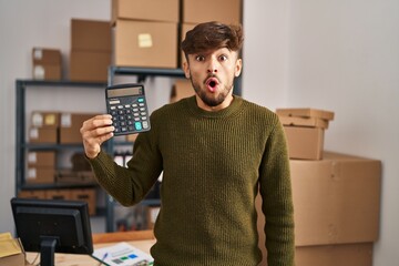 Arab man with beard working at small business ecommerce holding calculator scared and amazed with open mouth for surprise, disbelief face