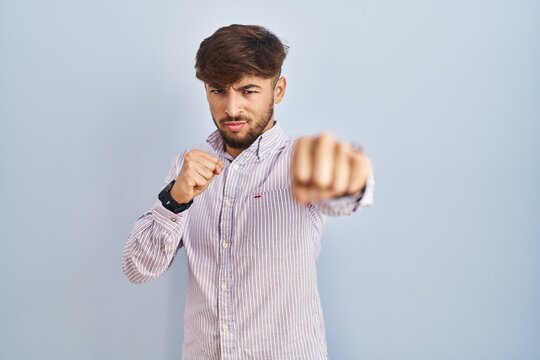 Arab man with beard standing over blue background punching fist to fight, aggressive and angry attack, threat and violence