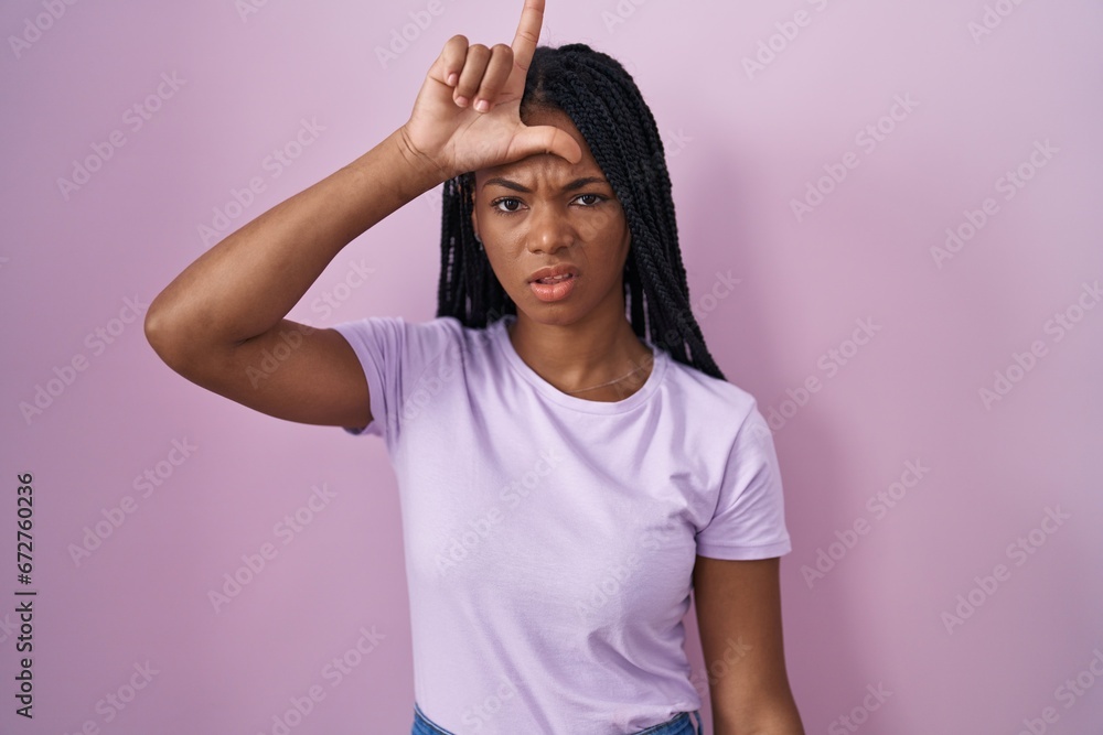 Wall mural African american woman with braids standing over pink background making fun of people with fingers on forehead doing loser gesture mocking and insulting.