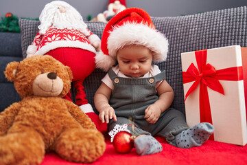 Fototapeta na wymiar Adorable hispanic toddler sitting on sofa wearing christmas hat at home