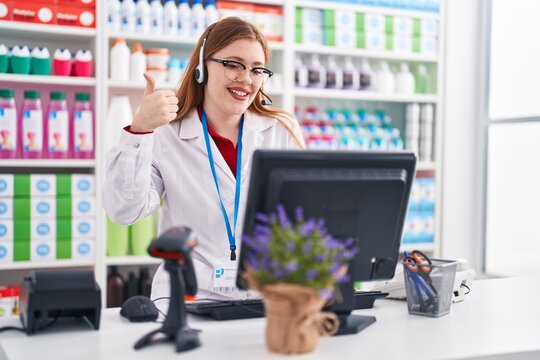 Redhead woman working at pharmacy drugstore wearing headset smiling happy and positive, thumb up doing excellent and approval sign