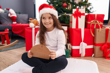 Obraz na płótnie Canvas Adorable hispanic girl holding santa claus letter sitting on floor by christmas tree at home