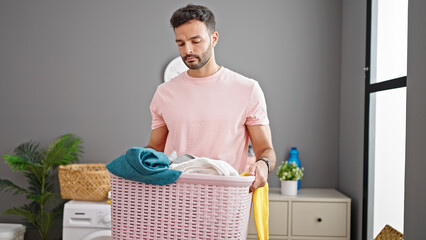 Young hispanic man holding basket with clothes at laundry room