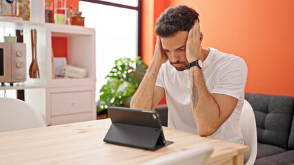 Young hispanic man using touchpad sitting on table stressed at dinning room