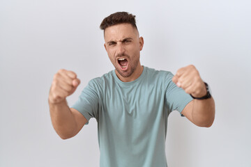 Hispanic man with beard standing over white background angry and mad raising fists frustrated and furious while shouting with anger. rage and aggressive concept.