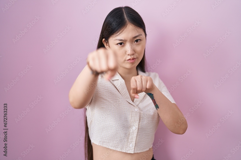 Sticker chinese young woman standing over pink background punching fist to fight, aggressive and angry attac