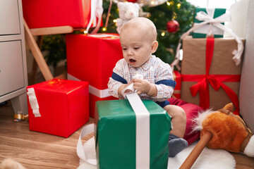 Fototapeta na wymiar Adorable caucasian baby unpacking gift sitting on floor by christmas tree at home