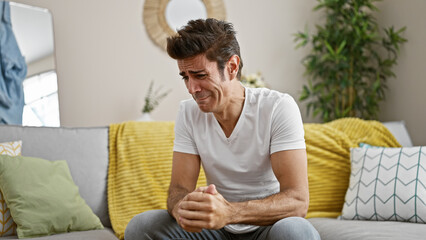 Young hispanic man stressed sitting on sofa crying at home