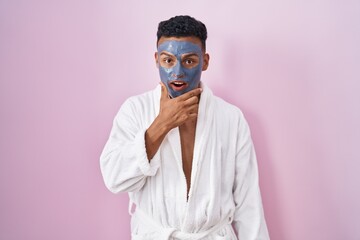Young hispanic man wearing beauty face mask and bath robe looking fascinated with disbelief, surprise and amazed expression with hands on chin