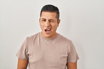 Hispanic young man standing over white background winking looking at the camera with sexy expression, cheerful and happy face.