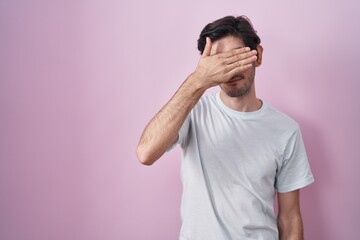 Young hispanic man standing over pink background covering eyes with hand, looking serious and sad....