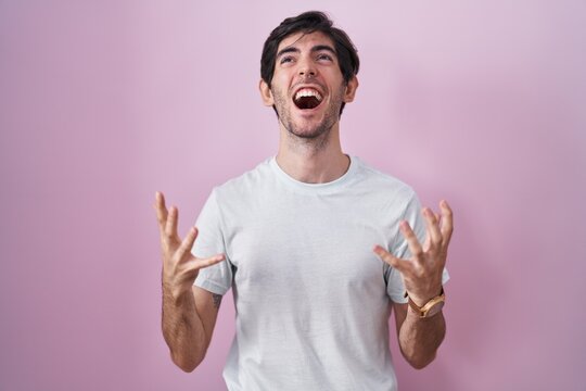 Young hispanic man standing over pink background crazy and mad shouting and yelling with aggressive expression and arms raised. frustration concept.
