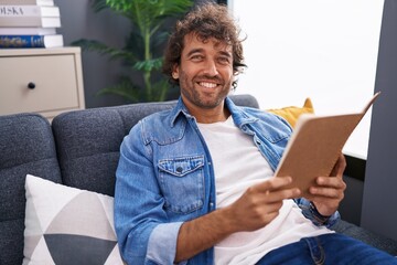 Young hispanic man reading book sitting on sofa at home - obrazy, fototapety, plakaty