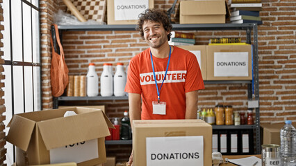 Young hispanic man volunteer smiling confident standing by donations cardboard box at charity center