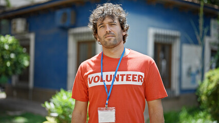 Young hispanic man activist wearing volunteer uniform standing with serious face at park