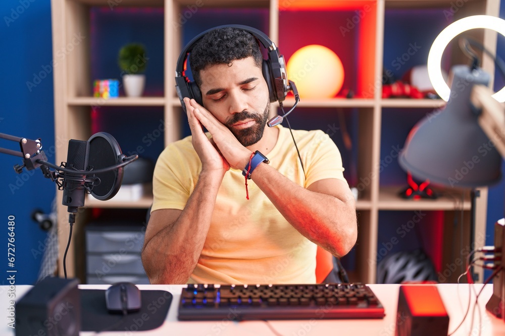 Poster Hispanic man with beard playing video games with headphones sleeping tired dreaming and posing with hands together while smiling with closed eyes.