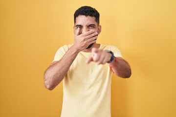 Hispanic man with beard standing over yellow background laughing at you, pointing finger to the...