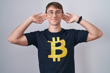 Caucasian blond man wearing bitcoin t shirt smiling pulling ears with fingers, funny gesture....