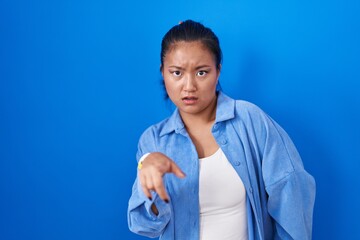 Asian young woman standing over blue background pointing displeased and frustrated to the camera,...