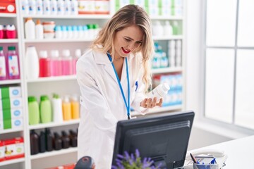 Young beautiful hispanic woman pharmacist using computer holding pills bottle at pharmacy