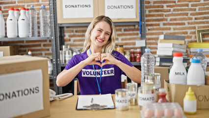 Young blonde woman volunteer sitting on table doing heart gesture at charity center