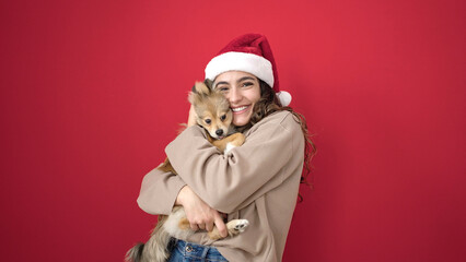 Fototapeta premium Young hispanic woman with dog smiling confident wearing christmas hat over isolated red background