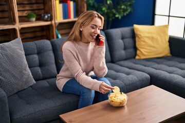 Young woman talking on smartphone eating chips potatoes at home