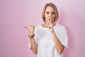 Young caucasian woman standing over pink background asking to be quiet with finger on lips pointing with hand to the side. silence and secret concept.