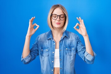 Young caucasian woman standing over blue background relax and smiling with eyes closed doing meditation gesture with fingers. yoga concept.