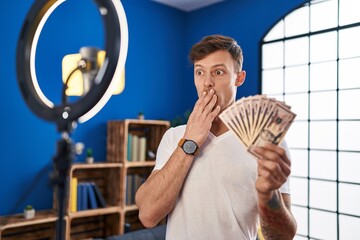Caucasian man recording vlog tutorial with smartphone at home holding money covering mouth with...