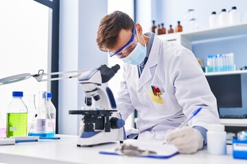 Naklejka na ściany i meble Young man scientist wearing medical mask using microscope writing document at laboratory