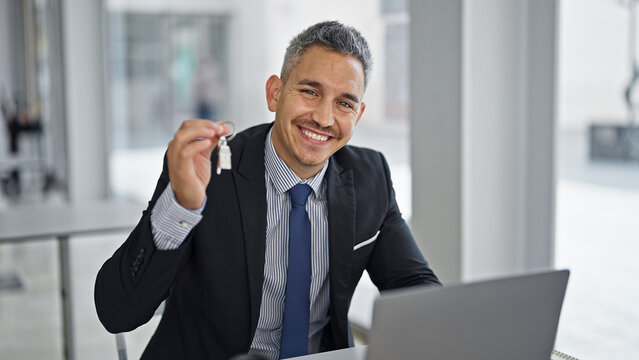 Young hispanic man real state agent holding new house keys using laptop at office