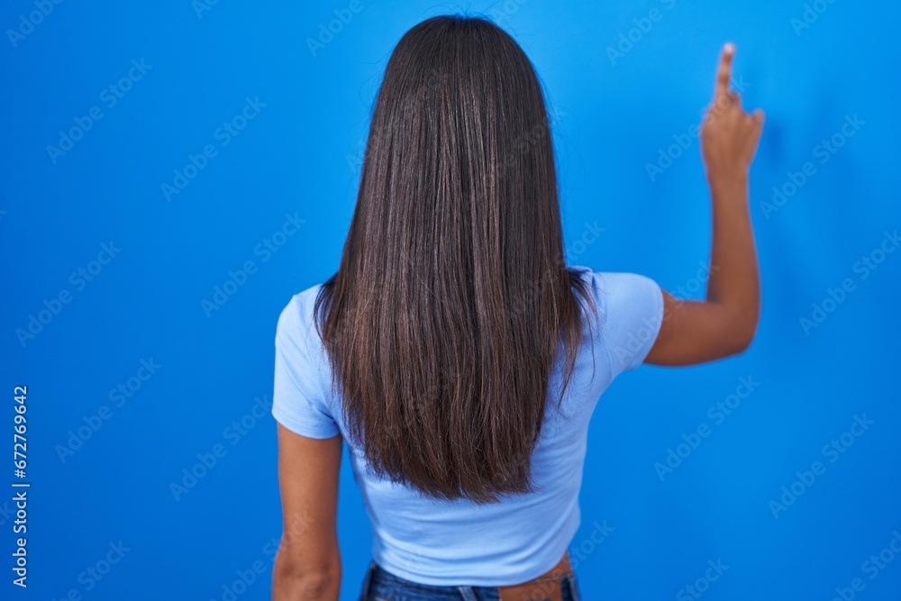 Wall mural Brunette young woman standing over blue background posing backwards pointing ahead with finger hand