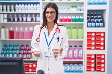 Young beautiful hispanic woman pharmacist holding pills and glass of water at pharmacy