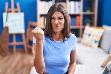 Brunette young woman sitting on the sofa at home angry and mad raising fist frustrated and furious while shouting with anger. rage and aggressive concept.