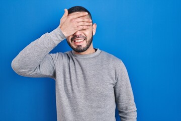 Hispanic man standing over blue background smiling and laughing with hand on face covering eyes for surprise. blind concept.