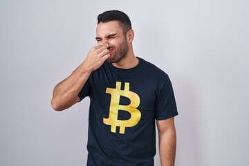Young hispanic man wearing bitcoin t shirt smelling something stinky and disgusting, intolerable smell, holding breath with fingers on nose. bad smell