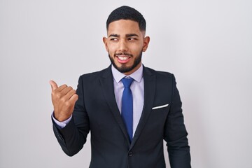 Young hispanic man wearing business suit and tie smiling with happy face looking and pointing to the side with thumb up.