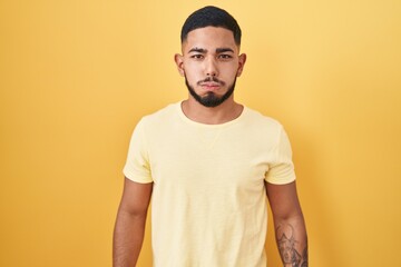 Young hispanic man standing over yellow background puffing cheeks with funny face. mouth inflated with air, crazy expression.