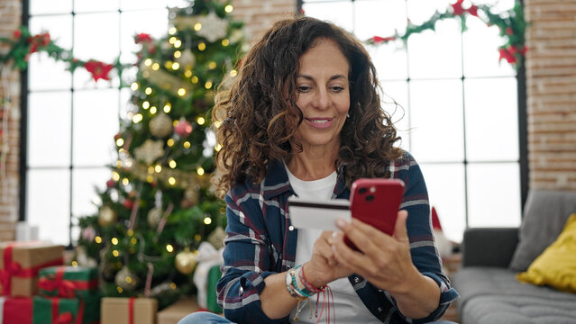 Middle age hispanic woman shopping with smartphone and credit card celebrating christmas at home