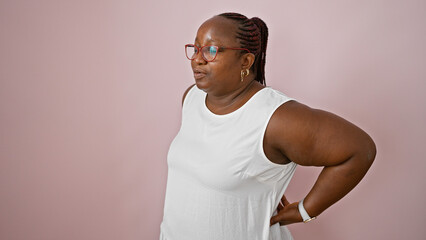 Worried african american woman, plus size with braids, standing alone, suffering serious backache over isolated pink background. worry of spinal injury showing on her hurting face. - obrazy, fototapety, plakaty