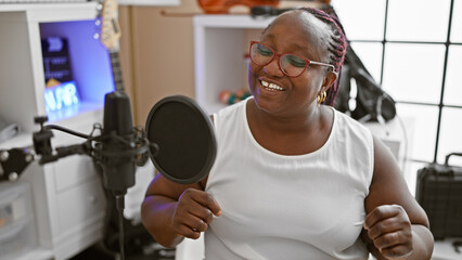 Confident african american woman, radiant smile, singing her song with passion at music studio,...