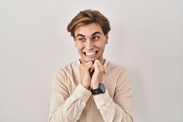 Young man standing over isolated background laughing nervous and excited with hands on chin looking...