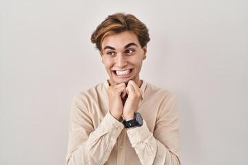 Young man standing over isolated background laughing nervous and excited with hands on chin looking to the side