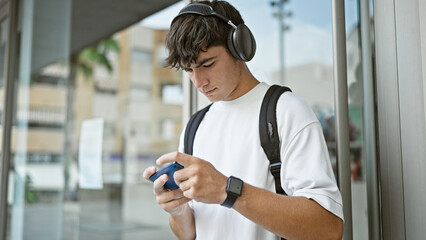 Young hispanic teenager student wearing backpack playing video game at university - obrazy, fototapety, plakaty