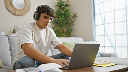Engrossed young hispanic male student chilling at home, sitting on comfy sofa, focused on studying online via laptop, perfectly relaxed, while seriously soaking up knowledge through headphones. - obrazy, fototapety, plakaty